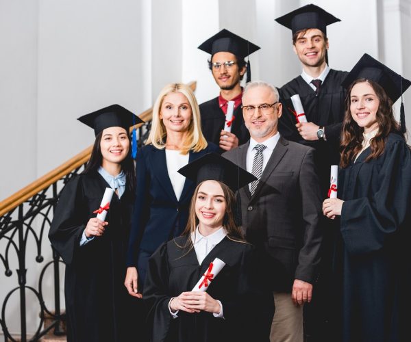 smiling students in graduation caps holding diplomas near happy teachers